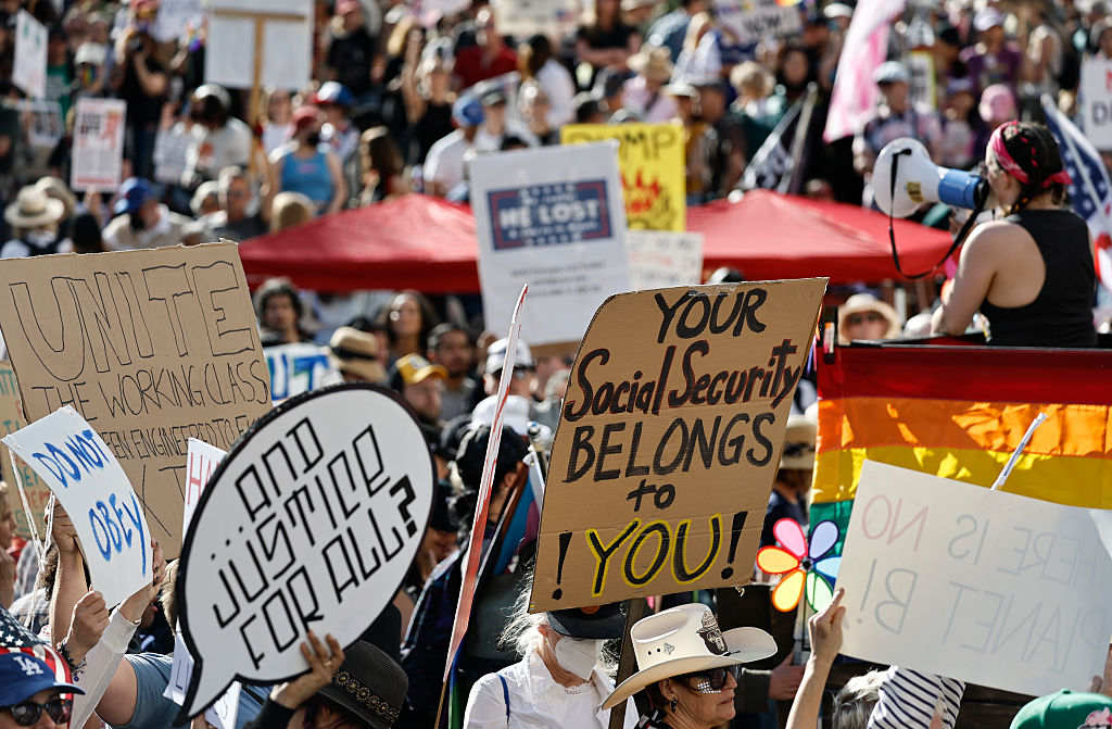 A large crowd of protesters holds up signs, including “Unite the working class,” “Your Social Security belongs to YOU!” and “Justice for All?” A person with a megaphone stands near a rainbow-colored flag.