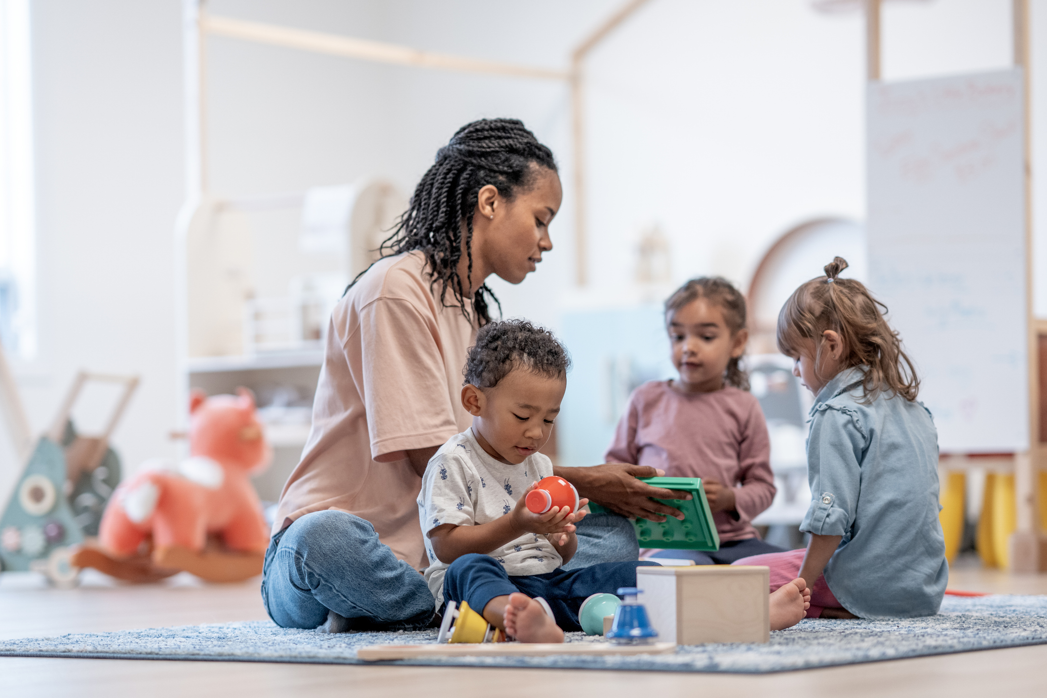A woman sits on the floor with three young children in a playroom, engaging them with toys and books. The background features shelves, a whiteboard, and a large stuffed animal.