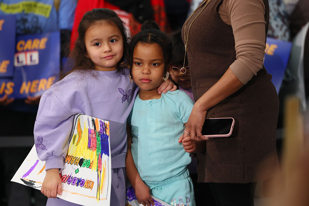 Two young girls stand close together, one holding a colorful drawing, while an adult beside them holds a phone. Both girls look toward the camera. People holding signs are visible in the background.
