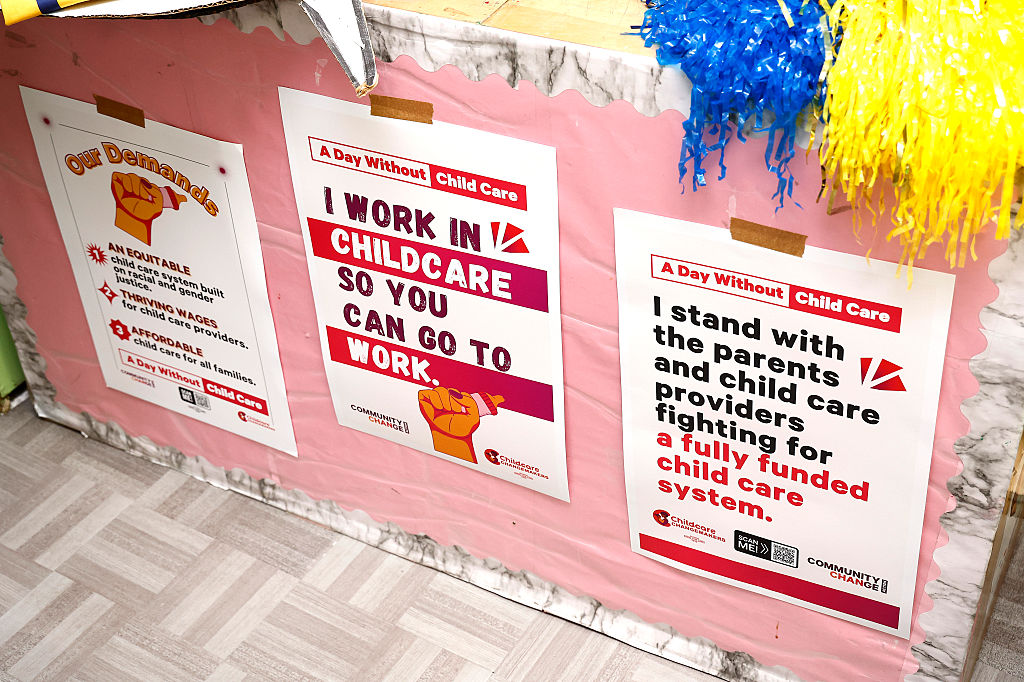Three posters on a pink wall advocate for child care support. Messages include: I work in childcare so you can go to work and I stand with parents and child care providers for a fully funded child care system.