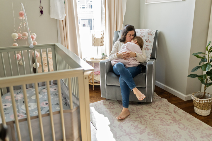 A woman sits in a gray armchair in a nursery, breastfeeding a baby. Sunlight streams through the window, illuminating a crib, soft rug, and a few decorative items in the cozy room.