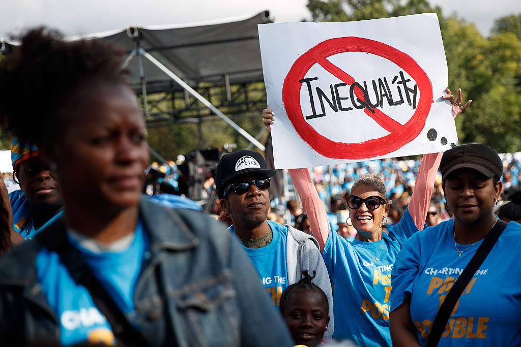 A smiling woman in a crowd holds a sign with Inequality crossed out by a red circle. People around her wear blue shirts at an outdoor event, advocating for equality.