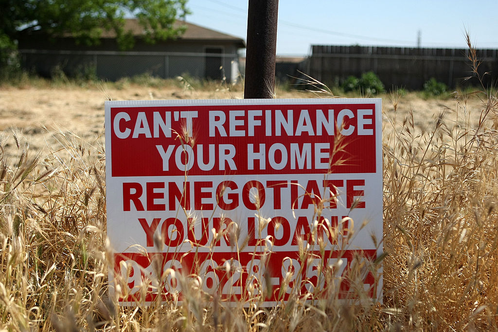A red and white sign in tall, dry grass reads CANT REFINANCE YOUR HOME, RENEGOTIATE YOUR LOAN with a phone number below. A house and a wooden fence are visible in the background.