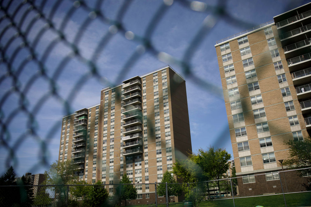 High-rise apartment buildings seen through a blurred chain-link fence, with trees and a blue sky in the background.