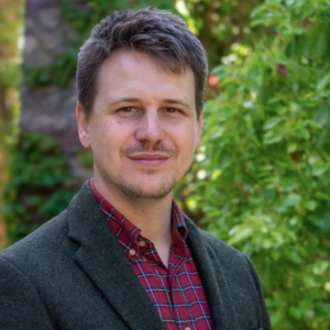 A man with short brown hair and slight stubble smiles outdoors, wearing a dark blazer over a red plaid shirt. Green trees and plants blur in the background.