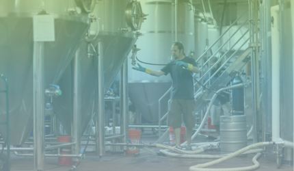 A person wearing gloves and boots works with large stainless steel brewing tanks in a brewery, with hoses and kegs on the floor.