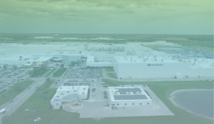 Aerial view of a large Subaru manufacturing facility surrounded by parking lots, green lawns, and a pond under a cloudy sky.