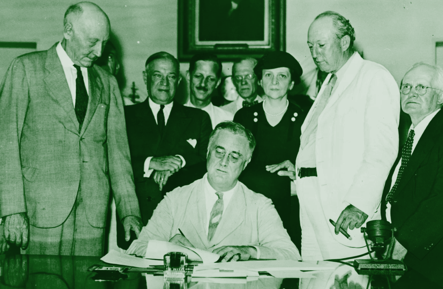 President Franklin D. Roosevelt sits at a desk signing a document, surrounded by several men and a woman, all dressed in formal attire, in a historical setting.