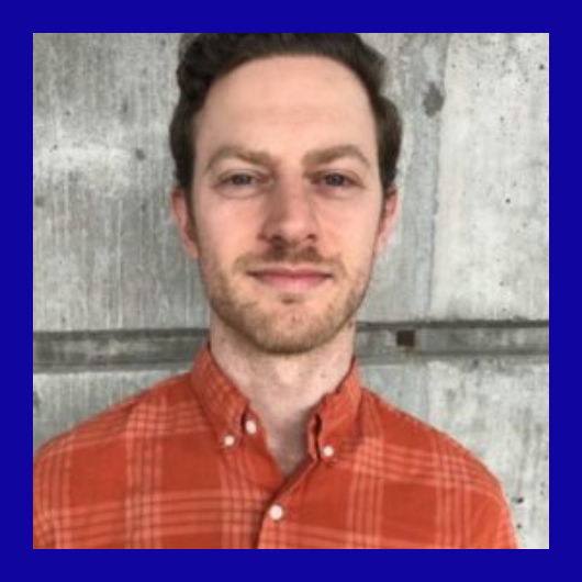 A man with short brown hair and a beard wearing an orange plaid shirt stands in front of a gray concrete wall. The image has a thick blue border.