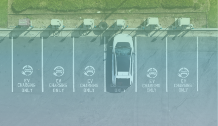 A white electric vehicle is parked and charging at one of several EV charging stations in a parking lot. The other charging spots are empty.