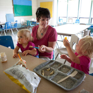 A woman sits at a table with two young children eating snacks in a bright, empty cafeteria. One child holds up food, while the other looks to the side. An empty meal tray and snack bags are on the table.