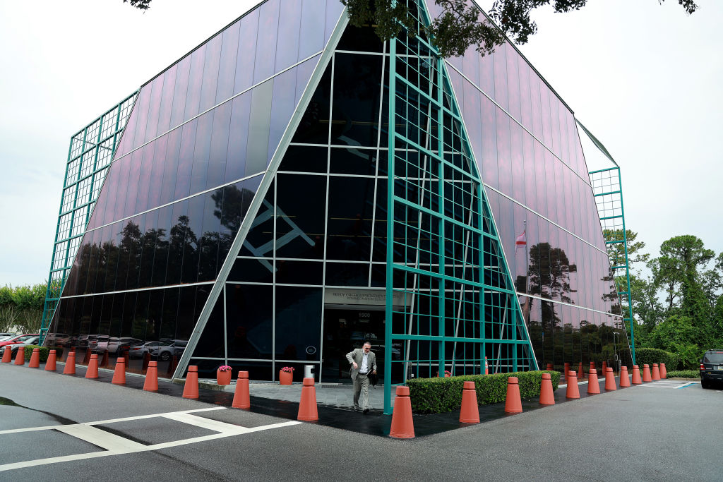 A modern glass office building with purple-tinted windows and teal frames. Orange traffic cones line the sidewalk, and a person walks out the front entrance. Trees and parked cars are visible around the building.