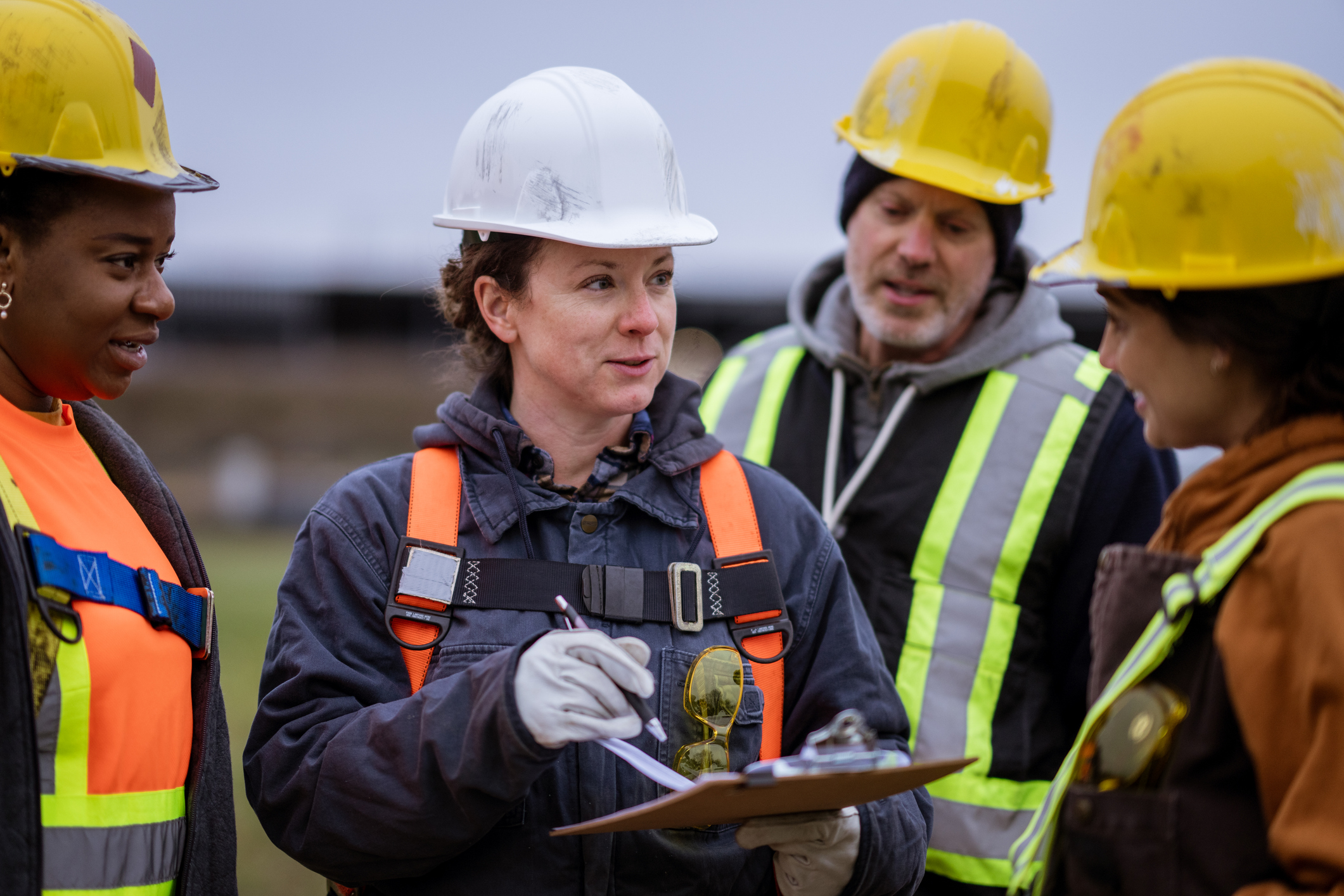 Four construction workers wearing safety gear and hard hats stand outdoors, gathered in a group. One person in the center holds a clipboard and pen, appearing to lead a discussion with the others.