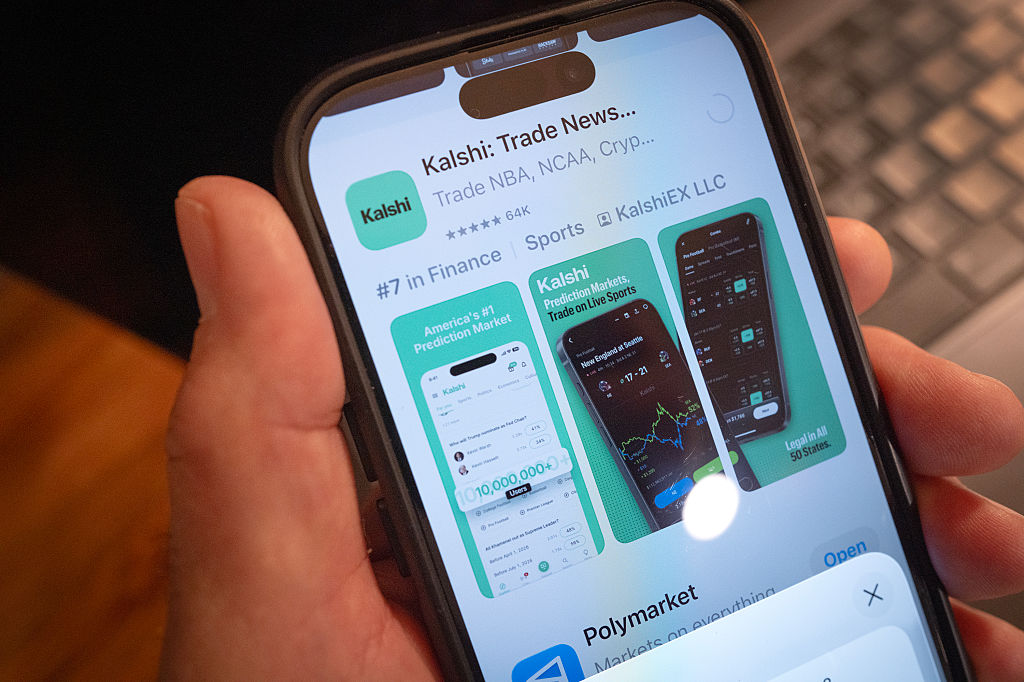 A hand holds a phone displaying the Kalshi app download page in the App Store, showing app details, rating, and preview images.