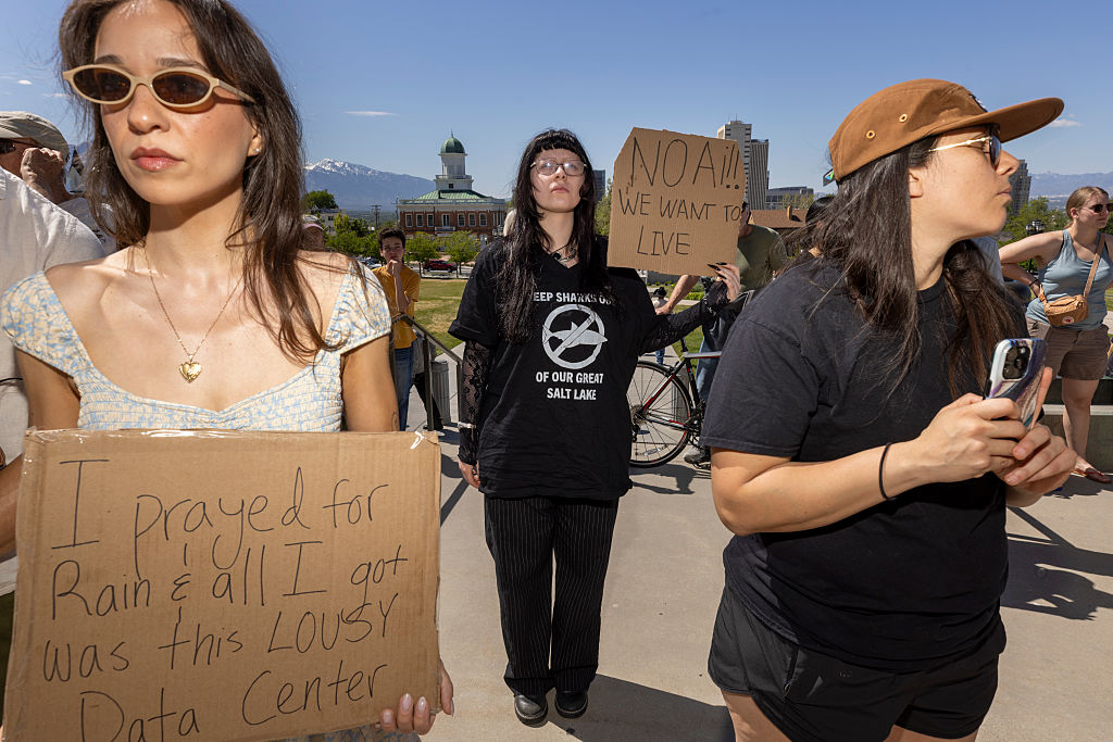 Three women at a protest hold cardboard signs. One reads, I prayed for rain & all I got was this LOUSY data center. Another sign says, NO!! WE WANT TO LIVE. People stand around under a sunny sky.