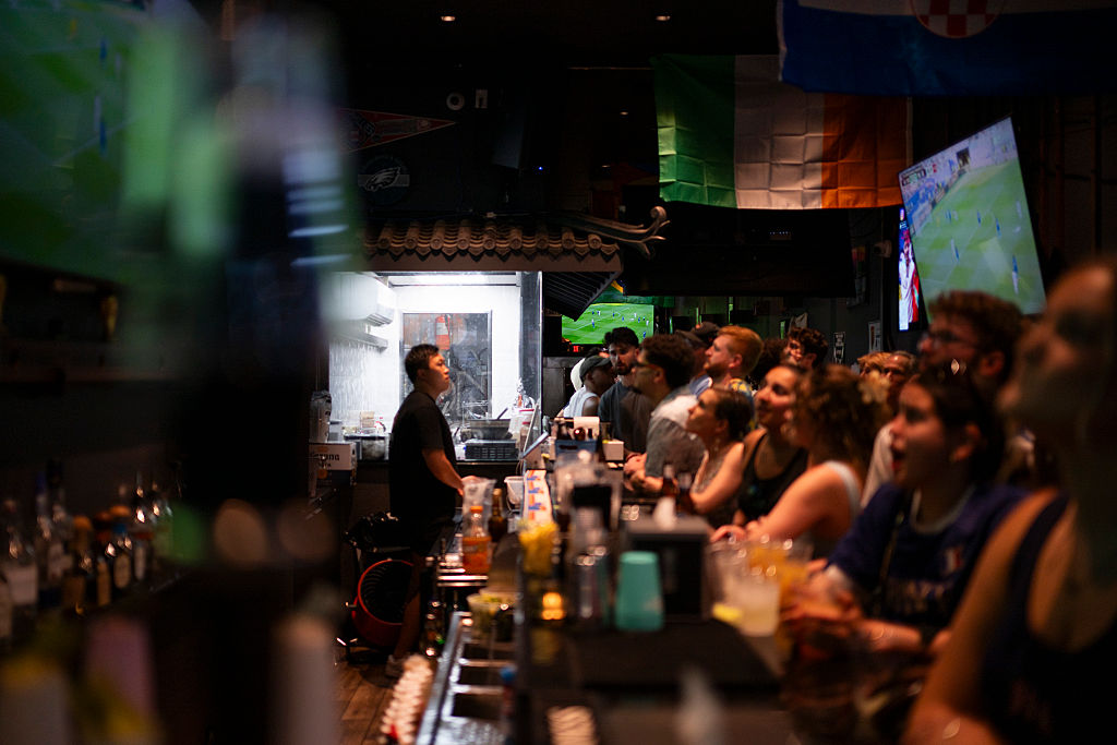 A group of people sit at a bar watching a soccer match on TV. The bartender stands behind the counter. Drinks are on the bar, and various flags hang from the ceiling. The scene is lively and energetic.