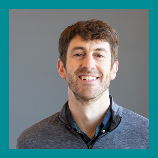 A smiling man with short brown hair and a beard, wearing a gray zip-up sweater over a blue shirt, stands in front of a plain gray background with a teal border around the image.