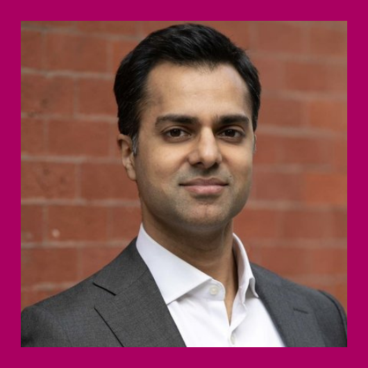 A man in a gray suit and white shirt stands in front of a brick wall, looking at the camera with a neutral expression. The image has a magenta border.