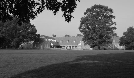 A large stone building with multiple chimneys is surrounded by trees and a grassy lawn. The photo is in black and white, with tree branches creating shadows in the foreground.