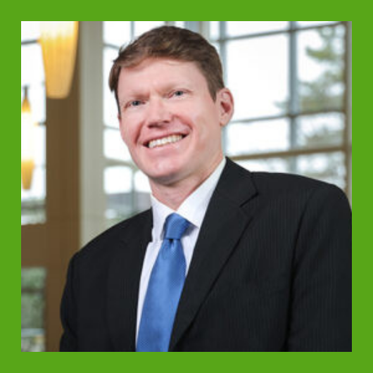 A smiling man in a black suit with a blue tie stands indoors near large windows. The photo has a green border.