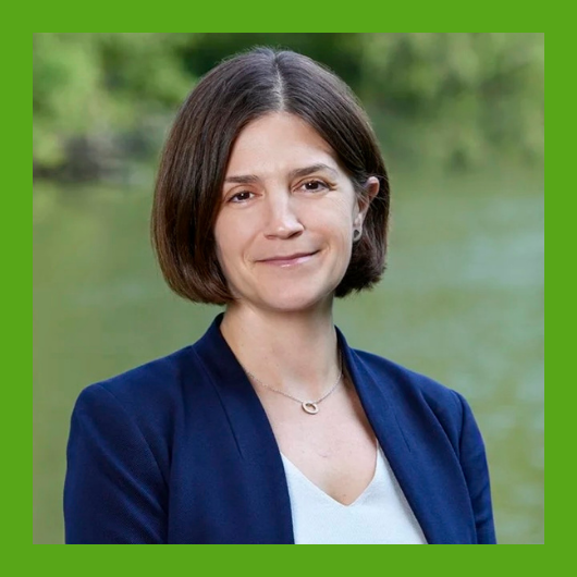 A woman with short brown hair, wearing a navy blazer and white top, stands outdoors with greenery and water in the background. The image has a green border.