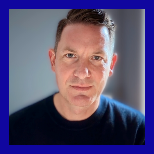 A man with short brown hair and light skin wearing a dark top, looking at the camera with a neutral expression. The background is blurred, and the image has a thick blue border.