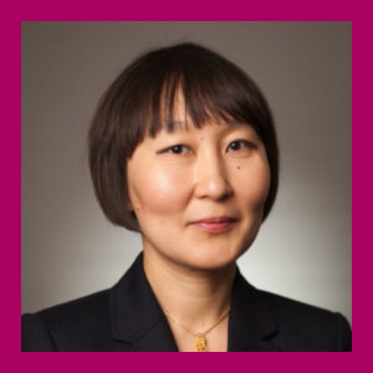 A woman with short dark hair wearing a black blazer and gold necklace poses for a professional headshot against a neutral background framed with a magenta border.