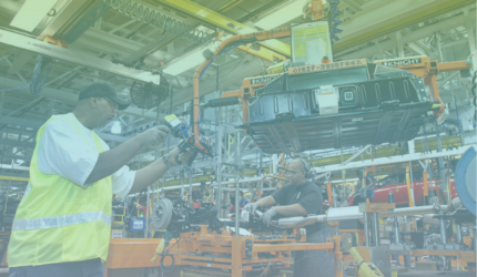 Two workers in safety gear assemble car parts in a brightly lit factory, operating machinery and tools surrounded by industrial equipment and suspended automotive components.