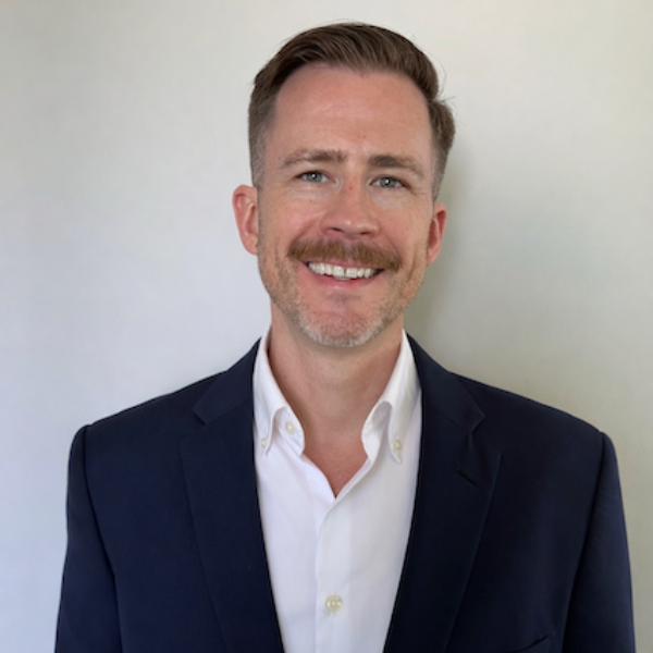 A smiling man with short light brown hair and a beard, wearing a navy suit jacket over a white dress shirt, stands in front of a plain light background.