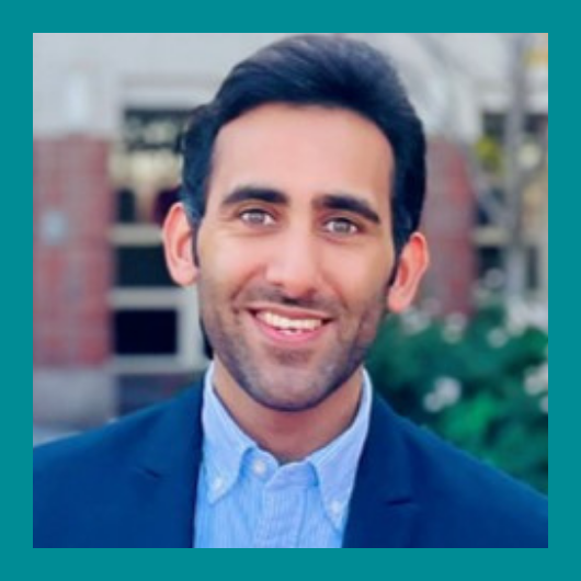 Smiling man with dark hair wearing a blue blazer and light blue shirt, standing outdoors in front of a building, with greenery and blurred background. The image has a teal border.