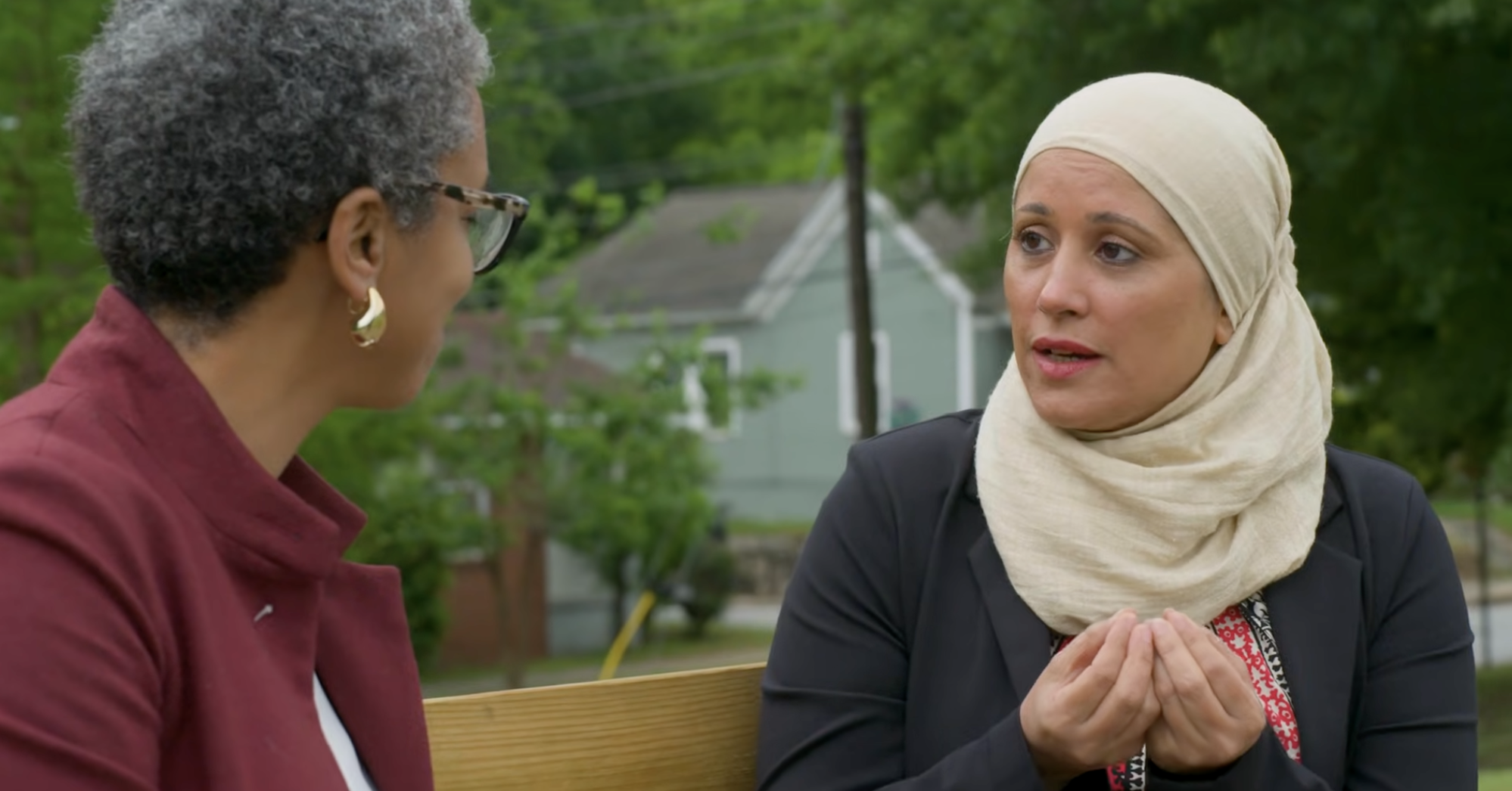Two women sit on a bench outdoors, engaged in conversation. One wears a cream hijab and gestures with her hands, while the other has short curly hair and glasses. Green trees and houses are visible in the background.