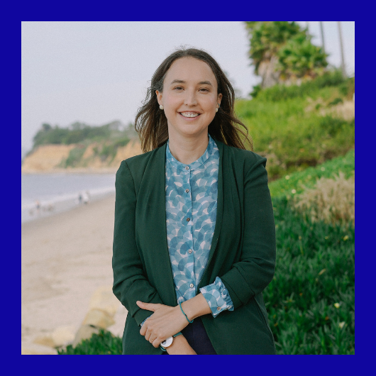 A woman stands on a beachside path, smiling, with greenery and cliffs in the background. She wears a green blazer over a patterned blue shirt, and the photo has a thick blue border.