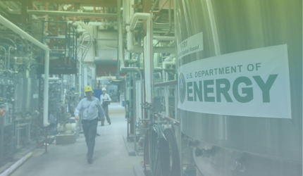 A large metal tank labeled “U.S. Department of Energy” inside an industrial facility, with two workers in hard hats walking amid pipes and equipment.