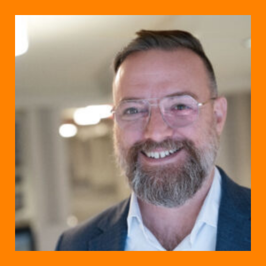 Smiling man with glasses, a beard, and short hair wearing a suit jacket and white shirt, standing indoors. The image has a bright orange border.