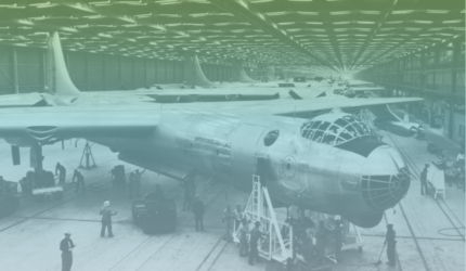 Vintage photograph of a large aircraft under construction in a hangar, surrounded by workers and equipment; multiple similar planes are visible in the background.