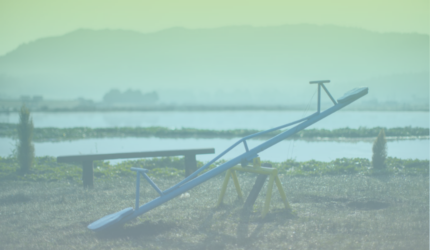 A blue seesaw stands on grassy ground near a wooden bench, with water, greenery, and distant hills visible in the background under a hazy sky.