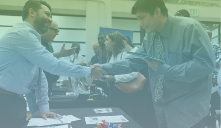 Two men shake hands across a table at a busy indoor event, possibly a job fair, with several people talking and exchanging documents in the background.