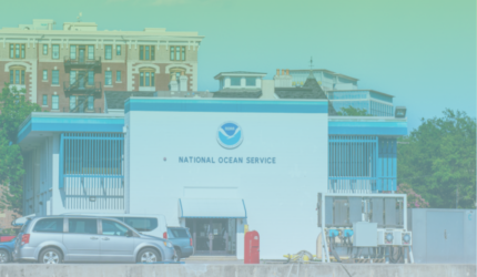 A building labeled “National Ocean Service” with cars parked in front; larger buildings and trees are visible in the background under a blue-green gradient sky.