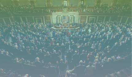 A large group of people stand in a legislative chamber with high ceilings and ornate decor, facing the front where a podium and flags are visible.