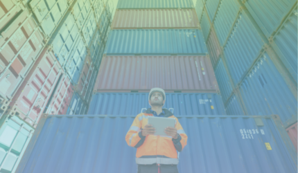 A worker in a safety helmet and orange jacket stands in front of stacked shipping containers, holding a tablet and looking up, possibly inspecting the containers at a cargo yard.