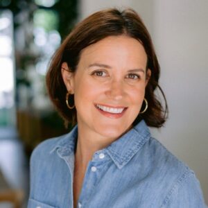 Woman with short brown hair and hoop earrings smiles at the camera, wearing a light blue denim shirt. The background is softly blurred, suggesting an indoor setting with natural light.