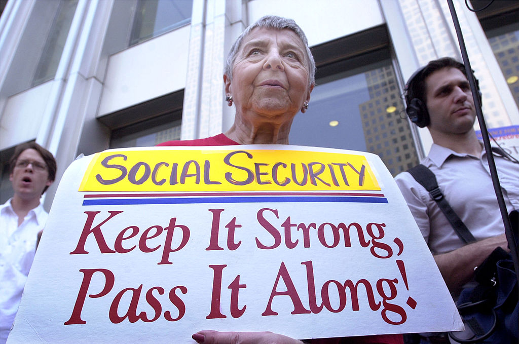 An older woman holds a sign reading Social Security: Keep It Strong, Pass It Along! at an outdoor event focused on social security, with two men standing behind her.