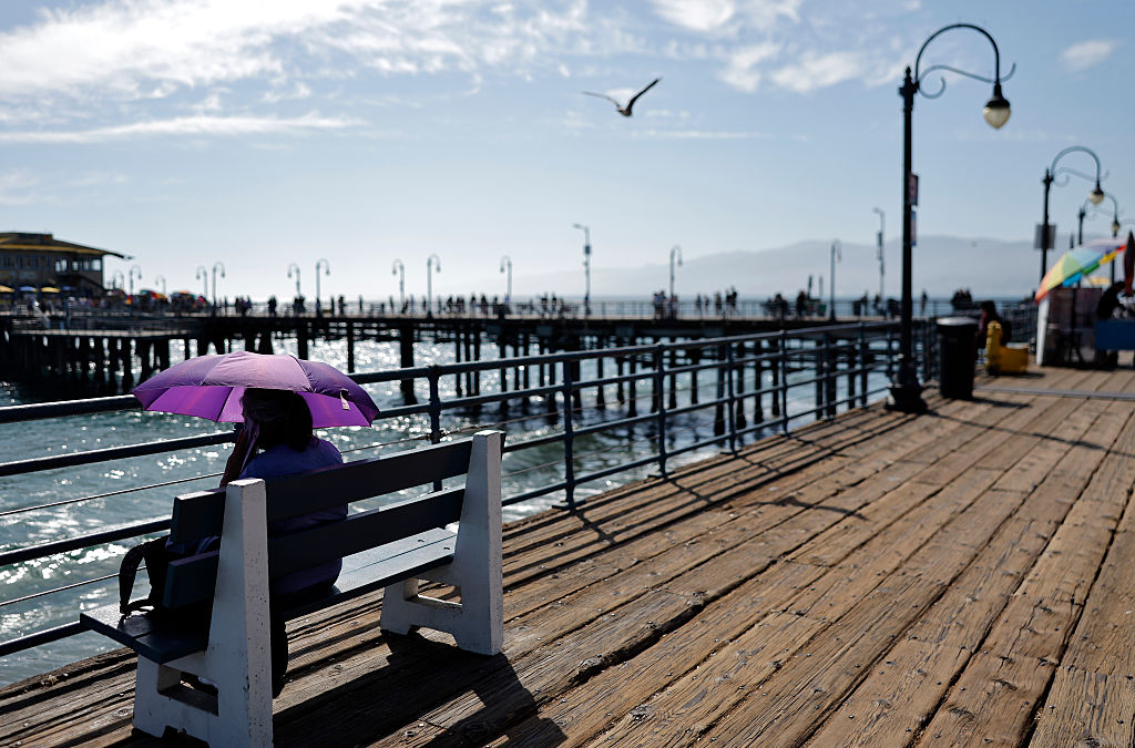 A person sits on a bench on a wooden pier, holding a purple umbrella. People walk in the distance. The sky is partly cloudy, and a bird flies overhead. Ocean waves are visible beside the pier.