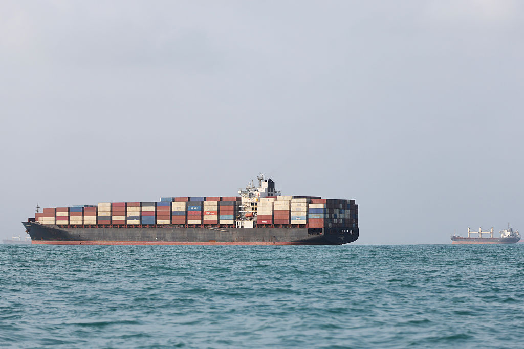 A large cargo ship loaded with colorful shipping containers sails on calm blue water, with another cargo vessel visible in the distance under a cloudy sky.