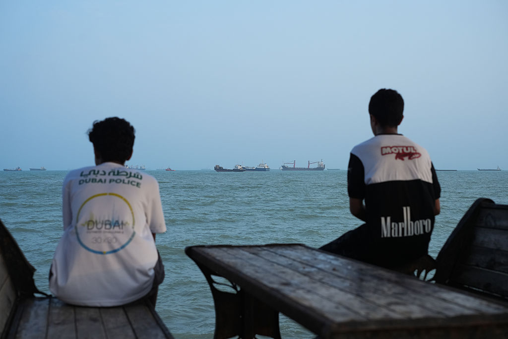 Two people sit on wooden benches facing the sea, watching several cargo ships in the distance under a clear sky. The scene appears calm and contemplative.