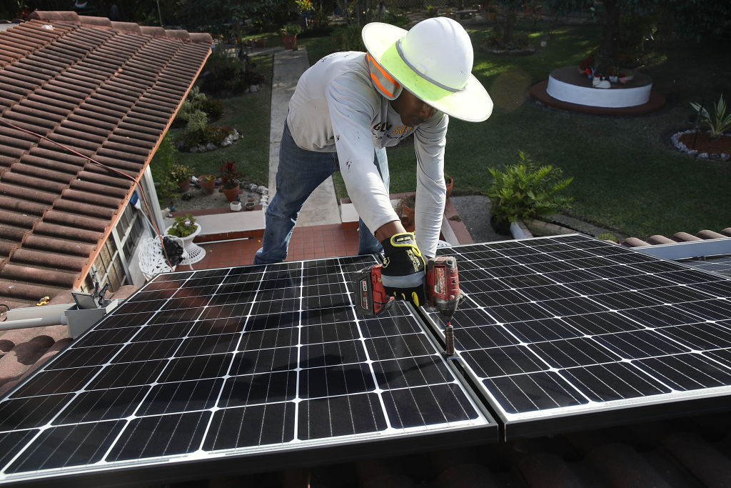 A worker wearing a wide-brim hat installs solar panels on a residential roof using a power drill, with a garden and trees visible in the background.