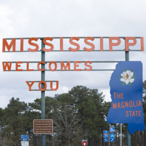 Large road sign reads Mississippi Welcomes You with a blue Magnolia State shape, a white magnolia flower, and trees in the background. Highway and informational signs are visible below. Cloudy sky above.