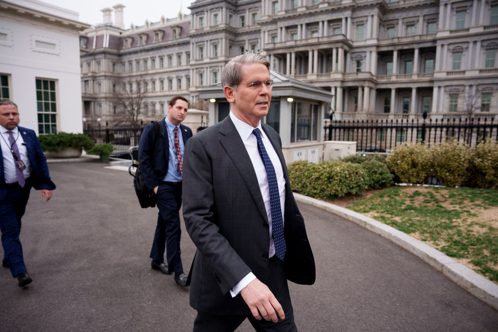 A man in a suit walks outside a government building, accompanied by two others in suits. The background features a large, ornate, multi-story structure with many windows and a black iron fence.