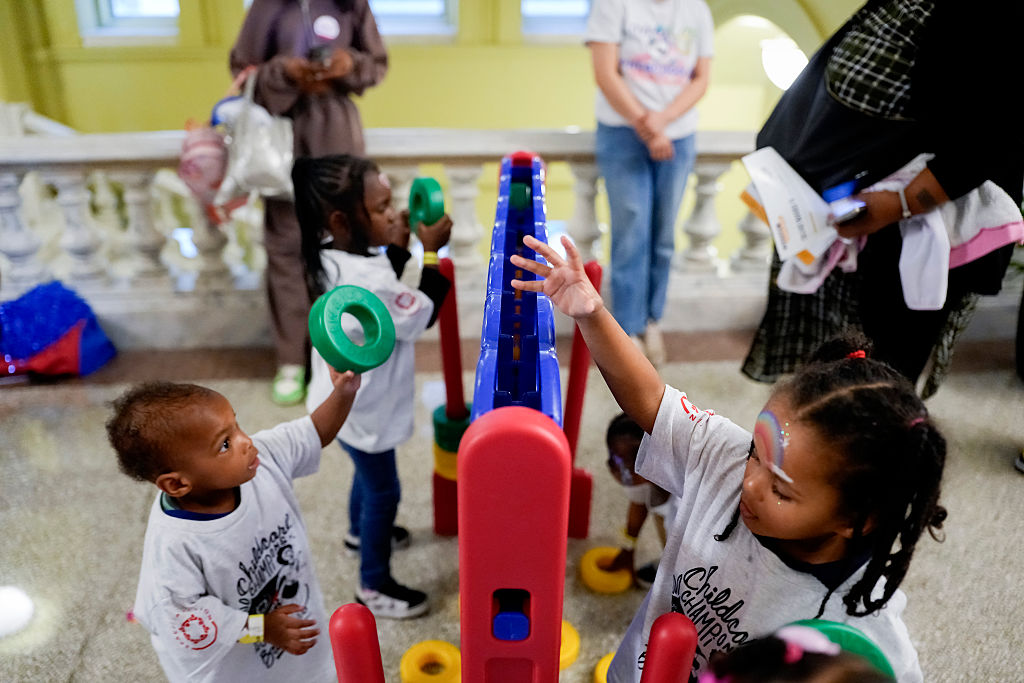 Several young children play with a giant Connect Four game indoors. They wear matching shirts, and some hold large plastic rings. Adults stand in the background, watching the kids.