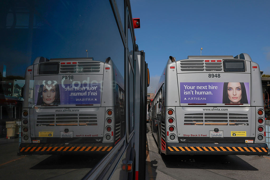 Two city buses parked side by side show ads with a womans face and the text Your next hire isnt human. The scene is reflected clearly in the window next to the buses.